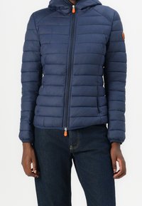 Person, der eine navyblaue Steppjacke mit Reißverschluss vorne und Kapuze trägt, kombiniert mit dunkelblauen Denim-Jeans, steht vor einem einfarbigen Hintergrund.