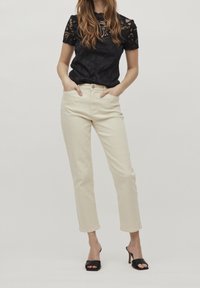 Donna in piedi con le mani in tasca, che indossa un top corto in pizzo nero, jeans beige a vita alta e sandali neri con tacco aperti.