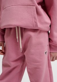Roze hoodie met een voorzak, gecombineerd met bijpassende roze sweatpants voorzien van witte trekkoorden en een klein decoratief logo.