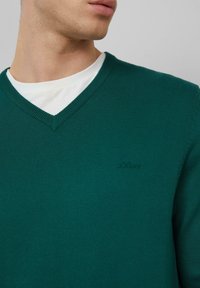 Grüner Strickpullover mit V-Ausschnitt, der ein Logo in passender Farbe auf der Brust und gerippte Details an den Bündchen aufweist.