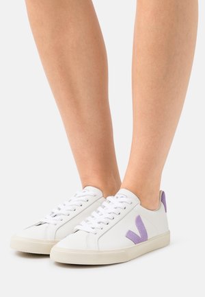 Bílé kožené tenisky s texturovaným fialovým V logem, plochými tkaničkami a béžovou gumovou podrážkou. Minimalistický design se zaměřením na čisté linie.
