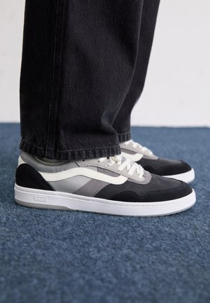 Černé a šedé semišové tenisky s bílými akcenty, s texturovaným povrchem, plochými tkaničkami a gumovou podrážkou. Na patě je logo značky.