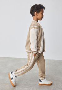 Tenue de sport beige comprenant une veste à boutons avec des manches blanches et un pantalon assorti avec des bandes latérales beiges et blanches. Baskets blanches.