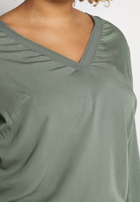 Top verde chiaro realizzato in morbido tessuto, con scollo a V e delicati dettagli di arricciatura sulle spalle e sui lati per un tocco di texture in più.