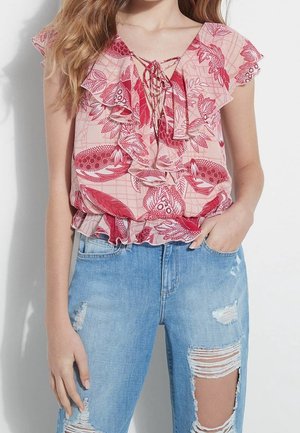 Femme portant un chemisier rose à volants avec des motifs floraux rouges et un jean bleu clair déchiré, main dans la poche.