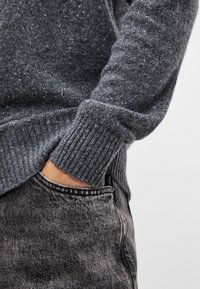 Pulover gri texturat cu manșete cu nervuri, asociat cu blugi din denim închis, cu o ușoară spălare și detalii la buzunarele din față.