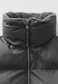 Giacca a vento verde scuro con colletto alto, dotata di una morbida fodera in pile grigio, chiusura frontale con zip e tirazip con il marchio. Tessuto liscio.