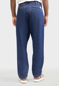 Džínsové nohavice z denimu v tmavomodrom prevedení, s voľným strihom, dvoma zadnými vreckami a viditeľnými švami. Nosené s béžovými teniskami.