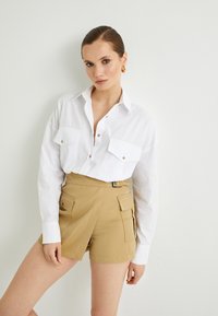 Camisa blanca de manga larga con botones dorados y dos bolsillos en el pecho, combinada con pantalones cortos de talle alto color caqui con cinturón y bolsillos laterales.