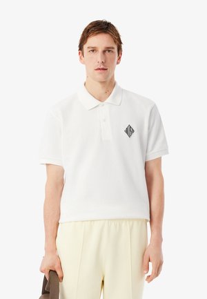 Witte polo met een zwart geometrisch logo op de linkerborst, voorzien van een klassieke kraag en korte mouwen. Gladde textuur.