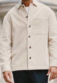 Mann in beiger Cordjacke mit Knöpfen und Brusttasche sowie dunklen Hosen, steht im Freien.