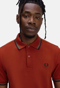 Rode polo shirt met contrasterende groene en blauwe gestreepte kraag, een twee-knoppen sluiting en geborduurd logo op de linkerborst. Gestructureerde stof.