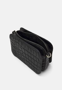 Borsa in pelle nera trapuntata con due scomparti con cerniera, appoggiata su uno sfondo bianco semplice. Presenta un anello metallico rotondo sul lato.