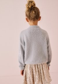 Maglione azzurro chiaro in morbido tessuto con maniche lunghe, abbinato a una gonna a strati in paillettes dorate lucide e soffici. Vista da dietro.