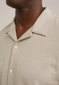 Camisa de manga corta beige con un patrón y una textura, con un cuello tipo spread y dos botones de color claro.