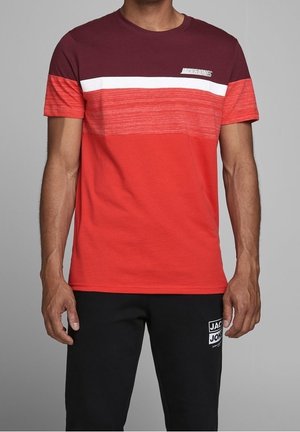 Homme portant un t-shirt à manches courtes à rayures rouges, bordeaux et blanches avec le logo "Jack & Jones" et un pantalon noir avec un logo sur la cuisse gauche.