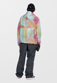 Kleurrijke, iriserende jas met een capuchon, voorzien van een draaiend patroon in roze, blauw en geel, gecombineerd met zwarte broek en handschoenen.