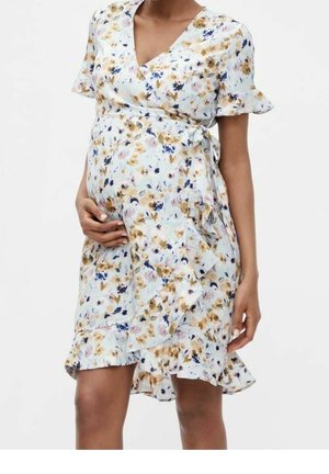 Pregnant woman wearing a white floral wrap dress with short ruffled sleeves and hand resting on her belly against a plain background.