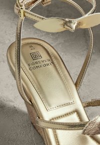 Zlatne metalne klizne sandale s teksturiranim uloškom i delikatnim trakama. Imaju dodatak u obliku lista na gornjem dijelu i oznaku "FOREVER COMFORT".