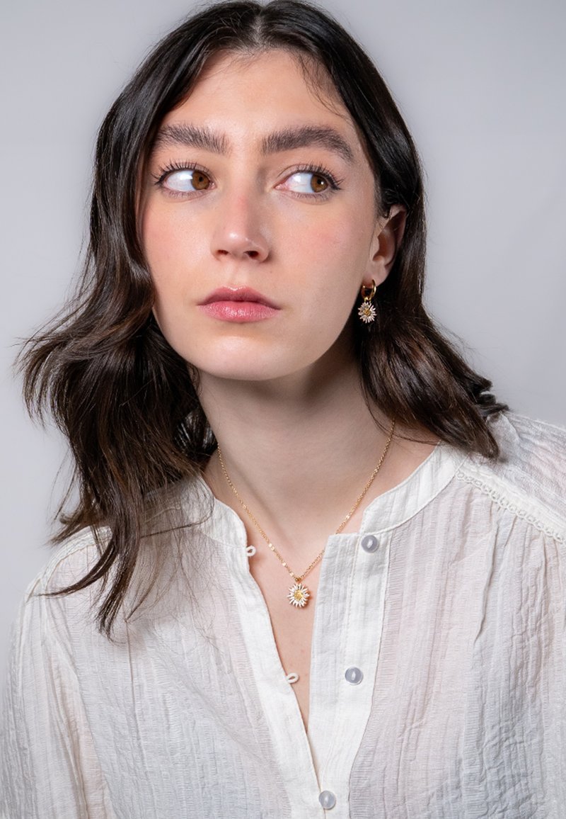 Guldsmycken med ett solstrålepandant och matchande örhängen, bärs tillsammans med en strukturerad vit blus. Mjukt, naturligt ljus framhäver detaljer.