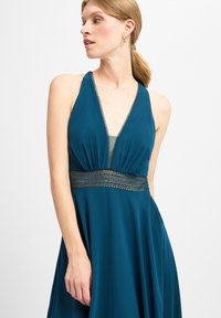 Teal-Halterkleid mit tiefem V-Ausschnitt, das ein strukturiertes Goldband in der Taille und fließenden Stoff für eine entspannte Silhouette aufweist.