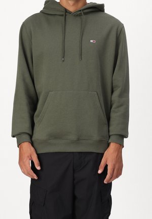 Mann trägt olivgrünen Hoodie mit Fronttasche und schwarze Cargohosen vor einfachem weißem Hintergrund.