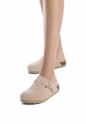 Zuecos de ante beige con punta cerrada, correa ajustable y acento de corcho en el tacón. Suela plana y texturizada para mayor agarre.