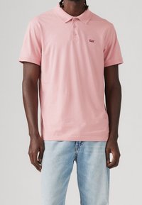 Polo rose en coton avec col, patte de boutonnage à trois boutons, manches courtes, et petit logo rouge sur la poitrine, associé à un jean bleu clair.