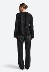 Juodas tekstūruotas švarkas su frakcinėmis detalėmis, ilgos rankovės ir apvaliu iškirpimu, derinamas su laisvai krentančiomis tamsiai pilkomis kelnėmis.