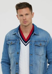 Veste en denim bleu clair avec deux poches poitrine et quincaillerie en cuivre, portée sur un pull en mailles torsadées blanc avec un col en V rouge et marine.