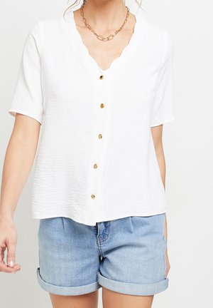 Vrouw draagt een witte blouse met korte mouwen en knopen met een getande halslijn en lichtblauwe opgerolde spijkershorts, staand tegen een effen achtergrond.