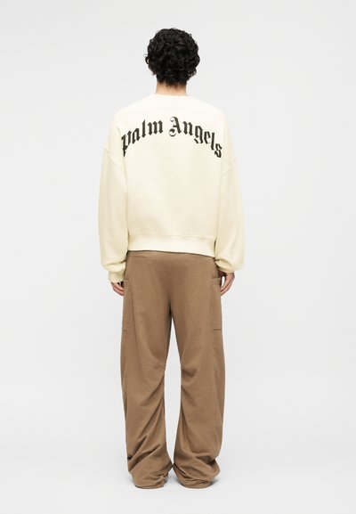 Άτομο με κοντά σγουρά μαλλιά φοράει κρεμ φούτερ με το κείμενο «Palm Angels» στην πλάτη και χαλαρό καφέ παντελόνι, να στέκεται μπροστά σε λευκό φόντο.