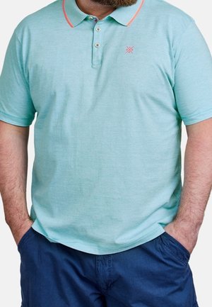 Mężczyzna ubrany w jasnoniebieską koszulkę polo z koralowym wykończeniem i granatowe krótkie spodenki, ręce w kieszeniach, zdjęcie przycięte poniżej barków.