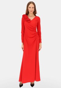 Długa czerwona sukienka z długimi rękawami, ozdobiona drapowanym wzorem oraz trójwymiarowym kwiatem na lewym ramieniu, gładka faktura materiału.