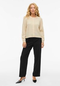 Cardigan beige clair avec un col rond, des manches longues et une fermeture à boutons sur le devant, associé à un pantalon noir taille haute et des escarpins noirs.