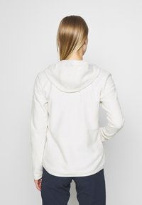 Frau steht mit dem Rücken zur Kamera, trägt eine weiße Kapuzenjacke und dunkle Hosen vor einem schlichten hellen Hintergrund.