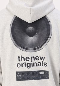 Lys grå hettegenser med et stort svart høyttaler graphic på baksiden, med svart tekst som leser "de nye originalene" og lydkontrolldials under.