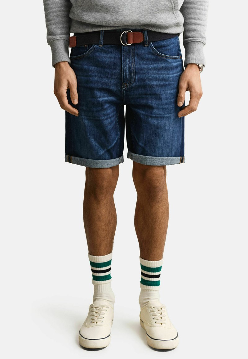 Homme portant un short en denim foncé roulé, un sweat-shirt gris, des chaussettes blanches rayées, des baskets blanches et une ceinture noire avec des accents en cuir marron.
