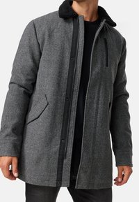 Grå uldblandingsfrakke med et herringbone mønster, sort kontrastlynlås og kant, samt tekstureret krave. Har sidelommer og en frontlynlås.