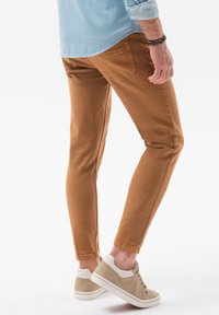 Pantalones de algodón de corte slim en color marrón, con una textura suave, que cuentan con bolsillos traseros y están combinados con zapatillas beige sobre un fondo claro.