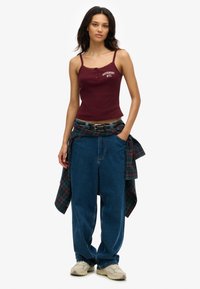 Burgundy ribbed tank top with lace trim and logo; paired with loose blue denim jeans and plaid shirt tied at the waist. Beige sneakers.