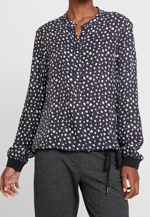 Person wearing a navy long-sleeve blouse with white irregular dots and dark gray pants, standing with arms relaxed.