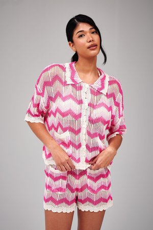 Jonge vrouw met donker haar draagt een roze-witte zigzag gehaakte top en bijpassende korte broek tegen een effen grijze achtergrond.