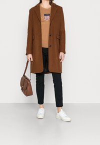 Brauner Wollmantel mit Schalkragen und drei Knöpfen, getragen über einem hellbraunen Sweatshirt und schwarzen, schmal geschnittenen Hosen, kombiniert mit weißen Sneakers und einer braunen Tasche.