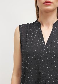 Černá bezramenní blůza s výstřihem do V, zdobená bílými puntíky a jemným leskem. Materiál vypadá hladce a je lehký.