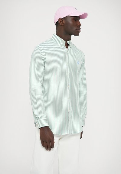 Zelena in bela črtasta srajca s srajčnim ovratnikom in logotipom na prsih, kombinirana z roza kapo in belimi hlačami.