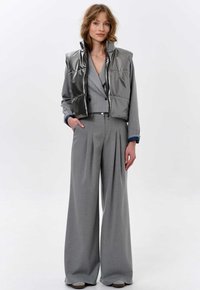 Metallisch silberne Steppweste über einem grauen Nadelstreifen-Blazer und weiten Hosen. Das Outfit hat einen lässigen Schnitt und sichtbare Nähte.