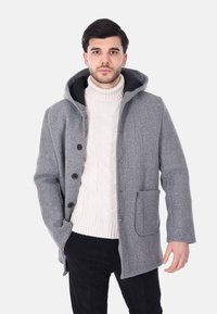 Cappotto grigio in lana con cappuccio, chiusura con bottoni e una tasca frontale. Abbinato a un maglione crema lavorato a trecce. Texture liscia.
