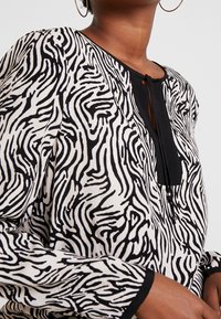 Maglietta a maniche lunghe con motivo a strisce zebra bianco e nero, caratterizzata da un collo rotondo, dettagli con bottoni e rifiniture nere lungo il collo e le maniche.
