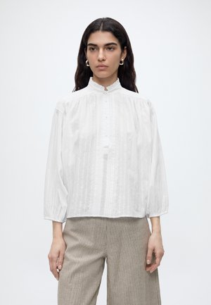 Femme aux longs cheveux foncés, portant une blouse blanche à manches longues avec des boutons et un pantalon beige à fines rayures, debout sur un fond uni.
